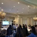 Attendees seated at round tables in an ornate room with chandeliers watch a speaker presenting at a podium beside a projected slide during the Alpha Omega Alpha Texas Chapter UTMB 2022 Fall Mixer.