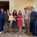 Group standing together in an ornate room during the Alpha Omega Alpha Texas Chapter UTMB 2022 Fall Mixer, with one attendee holding a framed certificate and two attendees holding a bouquet, posing near a podium.