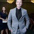 An individual in a gray suit stands smiling with hands in pockets in a ballroom at the Alpha Omega Alpha 2023 Induction Ceremony at UTMB, with another attendee holding papers and rows of chairs visible behind.