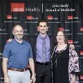 Three individuals pose together between red, white, and gray balloon columns at the Alpha Omega Alpha 2023 Induction Ceremony at UTMB, with a UTMB Health backdrop behind them.