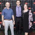 Three individuals pose for a full-length portrait at the Alpha Omega Alpha 2023 Induction Ceremony at UTMB, standing in front of a UTMB Health backdrop with balloon columns on both sides.