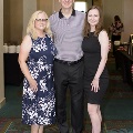 Three individuals pose together for a full-length group photo at the Alpha Omega Alpha 2023 Induction Ceremony at UTMB in a carpeted ballroom with tables and chairs in the background.