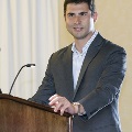An individual stands behind a wooden podium speaking into a microphone during the Alpha Omega Alpha 2023 Induction Ceremony at UTMB, with a curtain backdrop and presentation equipment visible.