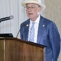 An individual stands behind a wooden podium speaking into a microphone during the Alpha Omega Alpha 2023 Induction Ceremony at UTMB, wearing a suit, hat, and patterned tie.
