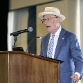 An individual wearing a light-colored hat stands at a wooden podium speaking into a microphone during the Alpha Omega Alpha 2023 Induction Ceremony at UTMB, with a projection screen and wall behind.