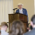 An individual stands at a podium speaking into a microphone during the Alpha Omega Alpha 2023 Induction Ceremony at UTMB, with audience members blurred in the foreground.