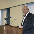 An individual stands behind a wooden podium speaking into a microphone during the Alpha Omega Alpha 2023 Induction Ceremony at UTMB, wearing a dark suit in a ballroom setting.