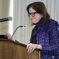 An individual stands at a wooden podium reading from notes during the Alpha Omega Alpha 2023 Induction Ceremony at UTMB, with a microphone and curtain backdrop in a ballroom setting.