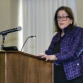 An individual speaks at a wooden podium with a microphone during the Alpha Omega Alpha 2023 Induction Ceremony at UTMB, wearing a patterned jacket and standing in a ballroom setting.