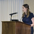 An individual stands at a wooden podium speaking into a microphone during the Alpha Omega Alpha 2023 Induction Ceremony at UTMB, with a projector and curtain backdrop behind.