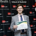 An individual poses holding a certificate at the Alpha Omega Alpha 2023 Induction Ceremony at UTMB, standing in front of a UTMB Health and John Sealy School of Medicine logo backdrop.