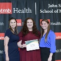 Three individuals stand together at the Alpha Omega Alpha 2023 Induction Ceremony at UTMB, with one holding a certificate in front of a UTMB Health and John Sealy School of Medicine branded backdrop.
