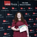 An individual stands between red, white, and gray balloon columns holding a certificate at the Alpha Omega Alpha 2023 Induction Ceremony at UTMB, with a UTMB Health and John Sealy School of Medicine backdrop.