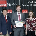 Three individuals stand together at the Alpha Omega Alpha 2023 Induction Ceremony at UTMB, with one holding a certificate in front of a UTMB Health and John Sealy School of Medicine logo backdrop.