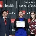 An individual stands centered holding a certificate during the Alpha Omega Alpha 2023 Induction Ceremony at UTMB, in front of a UTMB Health and John Sealy School of Medicine branded backdrop.