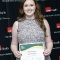 An individual holds a certificate reading “Congratulations Mary Fitzgibbon” at the Alpha Omega Alpha 2023 Induction Ceremony at UTMB, standing in front of a UTMB Health and John Sealy School of Medicine backdrop.