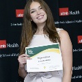 An individual holds a certificate reading “Congratulations Samantha Hobbs” at the Alpha Omega Alpha 2023 Induction Ceremony at UTMB, standing in front of a UTMB Health and John Sealy School of Medicine backdrop.
