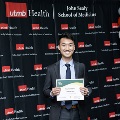 An individual stands centered between red, white, and gray balloon columns holding a certificate at the Alpha Omega Alpha 2023 Induction Ceremony at UTMB, with a UTMB Health backdrop.