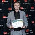 An individual stands holding a certificate at the Alpha Omega Alpha 2023 Induction Ceremony at UTMB, posing in front of a UTMB Health and John Sealy School of Medicine branded backdrop.