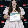 An individual holds a certificate and stands for a portrait at the Alpha Omega Alpha 2023 Induction Ceremony at UTMB, in front of a UTMB Health and John Sealy School of Medicine backdrop.