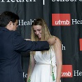 An individual places green honor cords around another individual’s neck during the Alpha Omega Alpha 2023 Induction Ceremony at UTMB, with a UTMB Health and John Sealy School of Medicine backdrop.