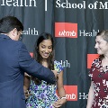 An individual places green honor cords around another individual’s neck during the Alpha Omega Alpha 2023 Induction Ceremony at UTMB, while another attendee stands nearby in front of a UTMB Health backdrop.