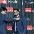 An individual places green honor cords around another individual’s neck during the Alpha Omega Alpha 2023 Induction Ceremony at UTMB, standing in front of a UTMB Health and John Sealy School of Medicine backdrop.