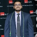 An individual stands for a portrait at the Alpha Omega Alpha 2023 Induction Ceremony at UTMB, wearing green honor cords and a suit in front of a UTMB Health and John Sealy School of Medicine backdrop with balloon decorations.