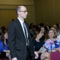 An individual in formal attire walks through a ballroom aisle during the Alpha Omega Alpha 2023 Induction Ceremony at UTMB, with seated attendees visible on both sides holding printed programs.