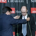 An individual places green honor cords around another individual’s neck during the Alpha Omega Alpha 2023 Induction Ceremony at UTMB, standing in front of a UTMB Health and John Sealy School of Medicine branded backdrop.