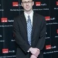 An individual stands for a formal portrait at the Alpha Omega Alpha 2023 Induction Ceremony at UTMB, wearing green honor cords in front of a UTMB Health and John Sealy School of Medicine branded backdrop.