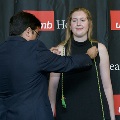 An individual places green honor cords around another individual’s neck during the Alpha Omega Alpha 2023 Induction Ceremony at UTMB, with a UTMB Health and John Sealy School of Medicine logo backdrop behind them.