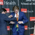 An individual places green honor cords around another individual’s neck during the Alpha Omega Alpha 2023 Induction Ceremony at UTMB, with a UTMB Health backdrop behind them.