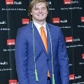 An individual stands in front of a UTMB Health and John Sealy School of Medicine backdrop, wearing green honor cords and a blue suit during the Alpha Omega Alpha 2023 Induction Ceremony.