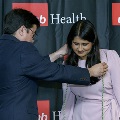 An individual places honor cords around another individual’s neck during the Alpha Omega Alpha 2023 Induction Ceremony at UTMB, with a UTMB Health and John Sealy School of Medicine backdrop behind them.