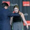 An individual places honor cords around another individual’s neck during the Alpha Omega Alpha 2023 Induction Ceremony at UTMB, with a UTMB Health and John Sealy School of Medicine backdrop behind them.