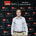 An individual stands centered between red, white, and gray balloon columns at the Alpha Omega Alpha 2023 Induction Ceremony at UTMB, wearing honor cords in front of a UTMB Health and John Sealy School of Medicine backdrop.