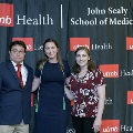 Three individuals stand together at the Alpha Omega Alpha 2023 Induction Ceremony at UTMB, posing in front of a UTMB Health and John Sealy School of Medicine backdrop with repeating logos.