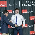 An individual places honor cords around another individual’s neck during the Alpha Omega Alpha 2023 Induction Ceremony at UTMB, with a UTMB Health and John Sealy School of Medicine backdrop behind them.