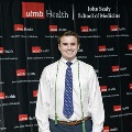 An individual poses at the Alpha Omega Alpha 2023 Induction Ceremony at UTMB, standing between red, white, and gray balloon columns with a UTMB Health and John Sealy School of Medicine backdrop behind.