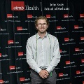 An individual stands centered between red, white, and gray balloon columns at the Alpha Omega Alpha 2023 Induction Ceremony at UTMB, wearing honor cords in front of a UTMB Health and John Sealy School of Medicine backdrop.