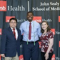 Three individuals pose together at the Alpha Omega Alpha 2023 Induction Ceremony at UTMB, standing in front of a UTMB Health and John Sealy School of Medicine logo backdrop.