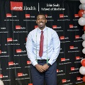 An individual stands centered between red, white, and gray balloon columns at the Alpha Omega Alpha 2023 Induction Ceremony at UTMB, wearing honor cords in front of a UTMB Health and John Sealy School of Medicine backdrop.