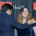An individual places honor cords around another individual’s neck during the Alpha Omega Alpha 2023 Induction Ceremony at UTMB, in front of a UTMB Health and John Sealy School of Medicine backdrop.