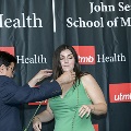 An individual places honor cords around another individual’s neck during the Alpha Omega Alpha 2023 Induction Ceremony at UTMB, in front of a UTMB Health and John Sealy School of Medicine backdrop.