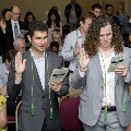 Individuals stand closely together in rows, raising one hand and reading from programs during the Alpha Omega Alpha 2023 Induction Ceremony at UTMB, with seated attendees and a formal event setting visible.