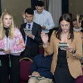 Several attendees stand in rows reading from printed programs and raising one hand during the Alpha Omega Alpha 2023 Induction Ceremony at UTMB, with seated guests visible in the foreground.