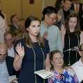 Several attendees stand together raising one hand and reading from printed programs during the Alpha Omega Alpha 2023 Induction Ceremony at UTMB, with seated guests visible behind them.
