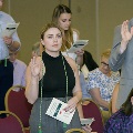 An individual stands holding a printed program while raising one hand during the Alpha Omega Alpha 2023 Induction Ceremony at UTMB, with other attendees participating nearby in a ballroom setting.