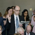 Several individuals stand closely together reading from printed programs during the Alpha Omega Alpha 2023 Induction Ceremony at UTMB, with seated attendees visible in the foreground and a decorated event space in the background.